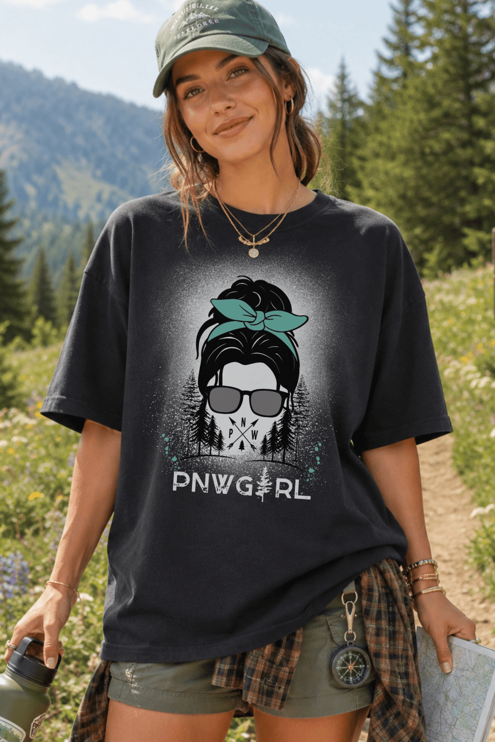 A woman wearing a dark charcoal graphic t-shirt with a 'PNWG GIRL' design, styled outdoors in a forest setting with an olive green cap, plaid shirt tied around her waist, and shorts.