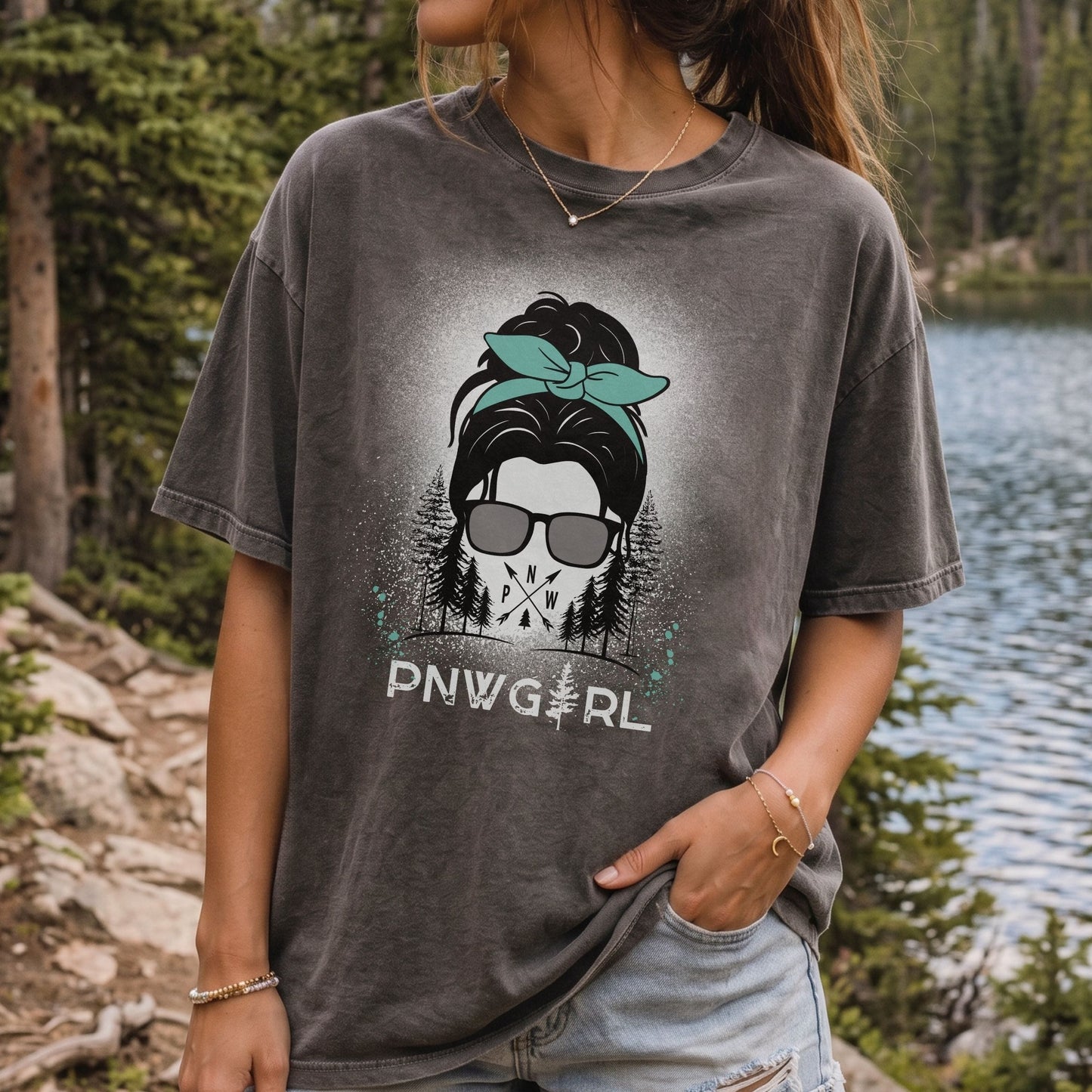 A woman wearing a faded charcoal gray graphic t-shirt with a 'PNWGirl' design, standing outdoors near a lake and pine trees.