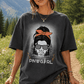 A woman outdoors on a mountain trail wearing an oversized dark charcoal graphic T-shirt with a distressed front print showing a woman in sunglasses and a red headscarf, plus the text 'DNWGRL'. She is also wearing an olive green cap, shorts, and a plaid shirt tied around her waist.