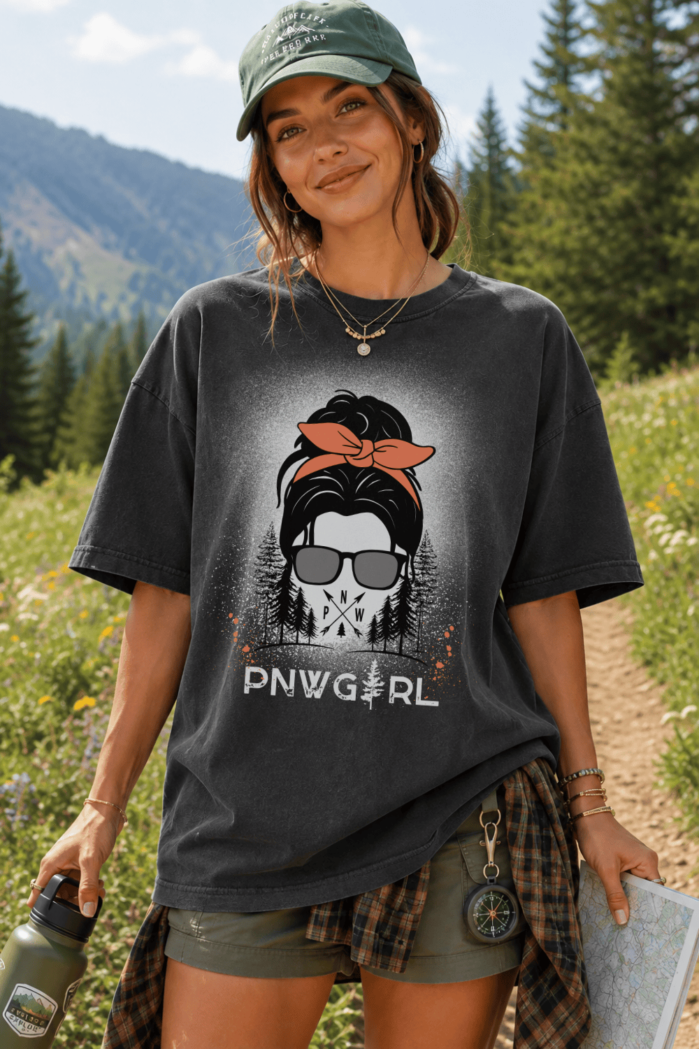 A woman outdoors on a mountain trail wearing an oversized dark charcoal graphic T-shirt with a distressed front print showing a woman in sunglasses and a red headscarf, plus the text 'DNWGRL'. She is also wearing an olive green cap, shorts, and a plaid shirt tied around her waist.
