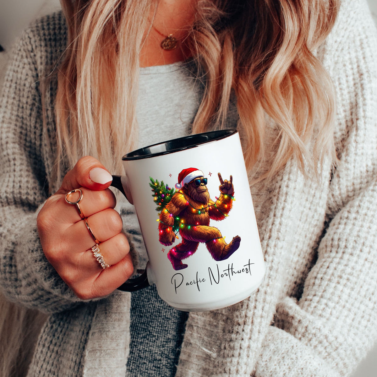 A person in a gray knit cardigan holds a white ceramic mug with a black handle and interior. The mug features a colorful illustration of a Santa-clad yeti carrying a Christmas tree, with the text 'Pacific Northwest' printed below.