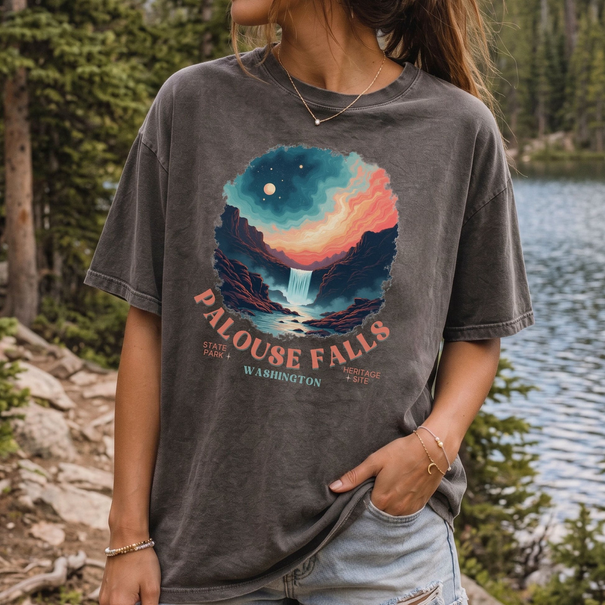 A woman wearing a faded charcoal gray graphic t-shirt featuring a colorful illustration of a waterfall and stars, with the text 'PALOUSE FALLS WASHINGTON' printed below. She is standing outdoors near a lake and pine trees.