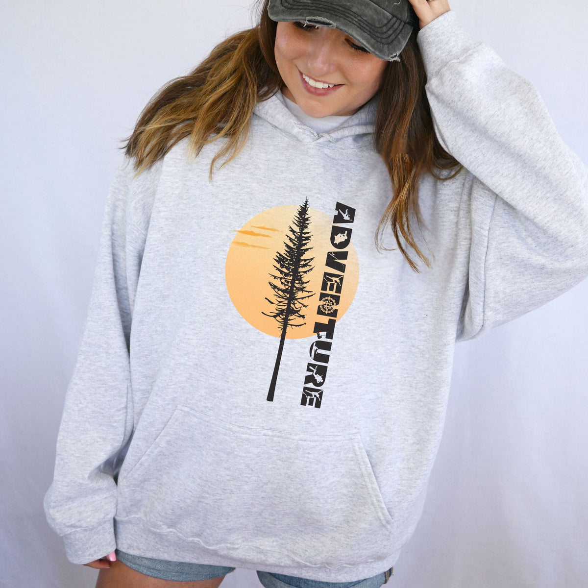 A woman wearing a light heather gray hoodie with a graphic print of a pine tree in front of an orange sun and the word 'ADVENTURE' in black, paired with a dark gray baseball cap.
