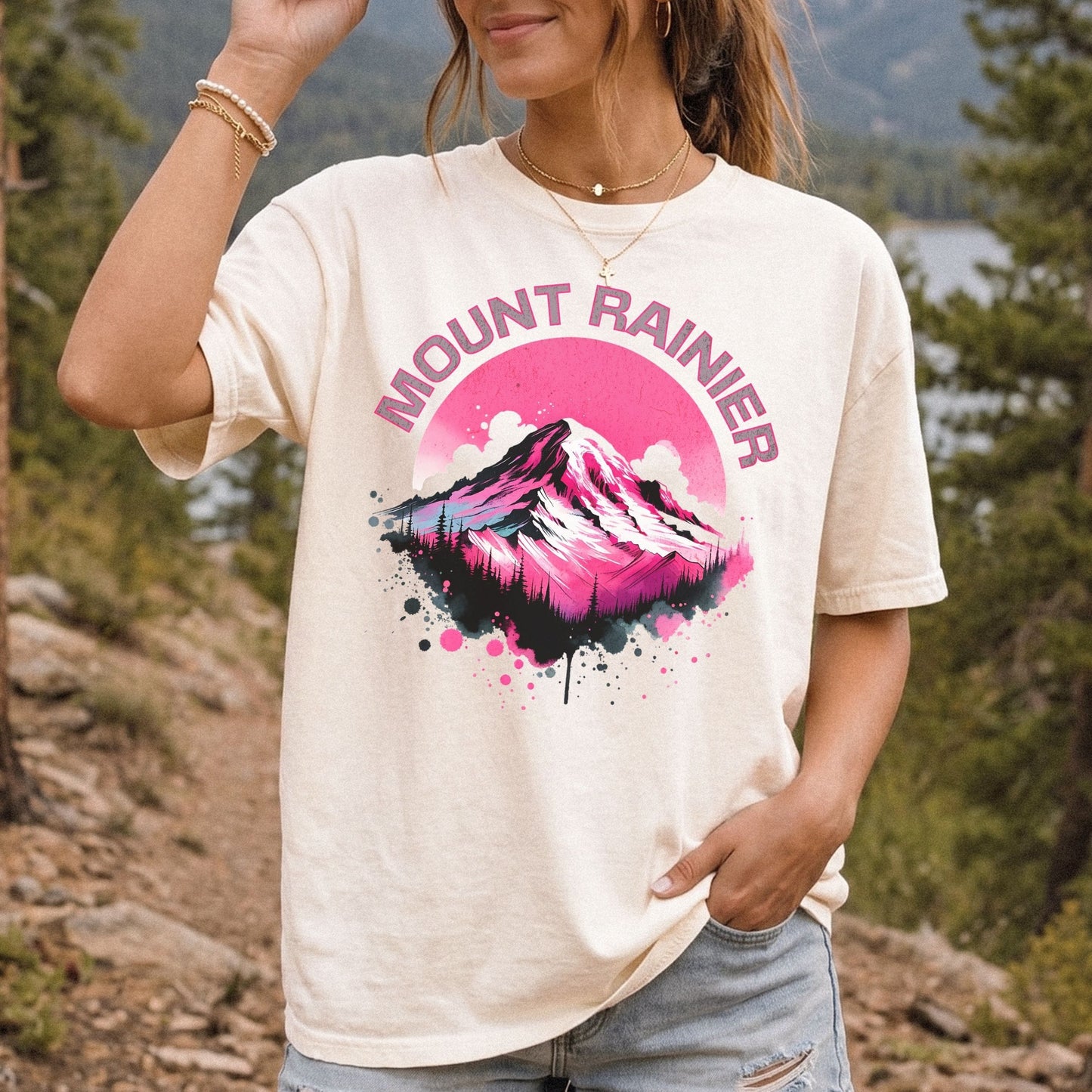 A woman wearing a cream-colored graphic t-shirt with a pink and black mountain design and the text 'MOUNT RAINIER' on the front, set against a blurred outdoor background of trees and a lake.