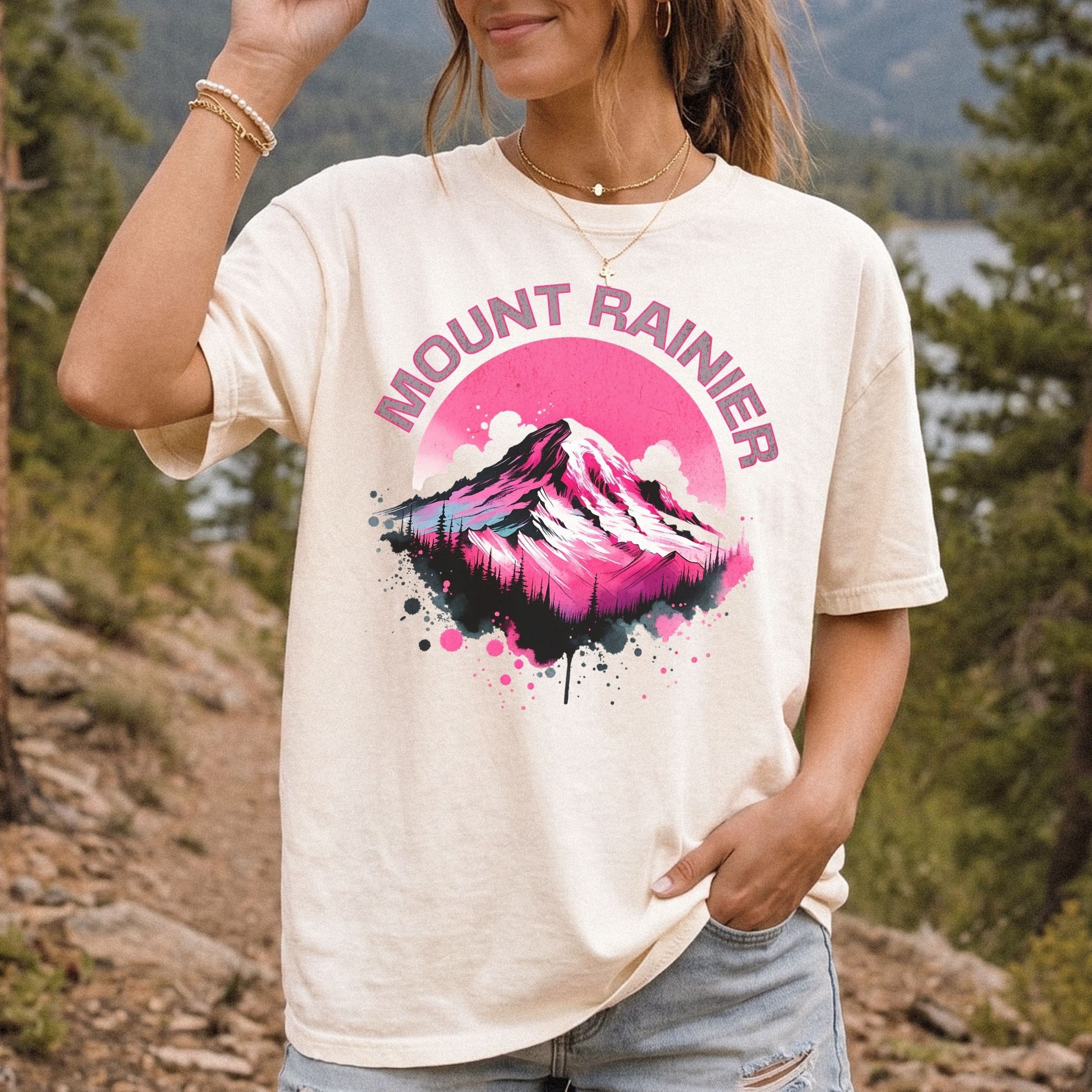 A woman wearing a cream-colored graphic t-shirt with a pink and black mountain design and the text 'MOUNT RAINIER' on the front, set against a blurred outdoor background of trees and a lake.