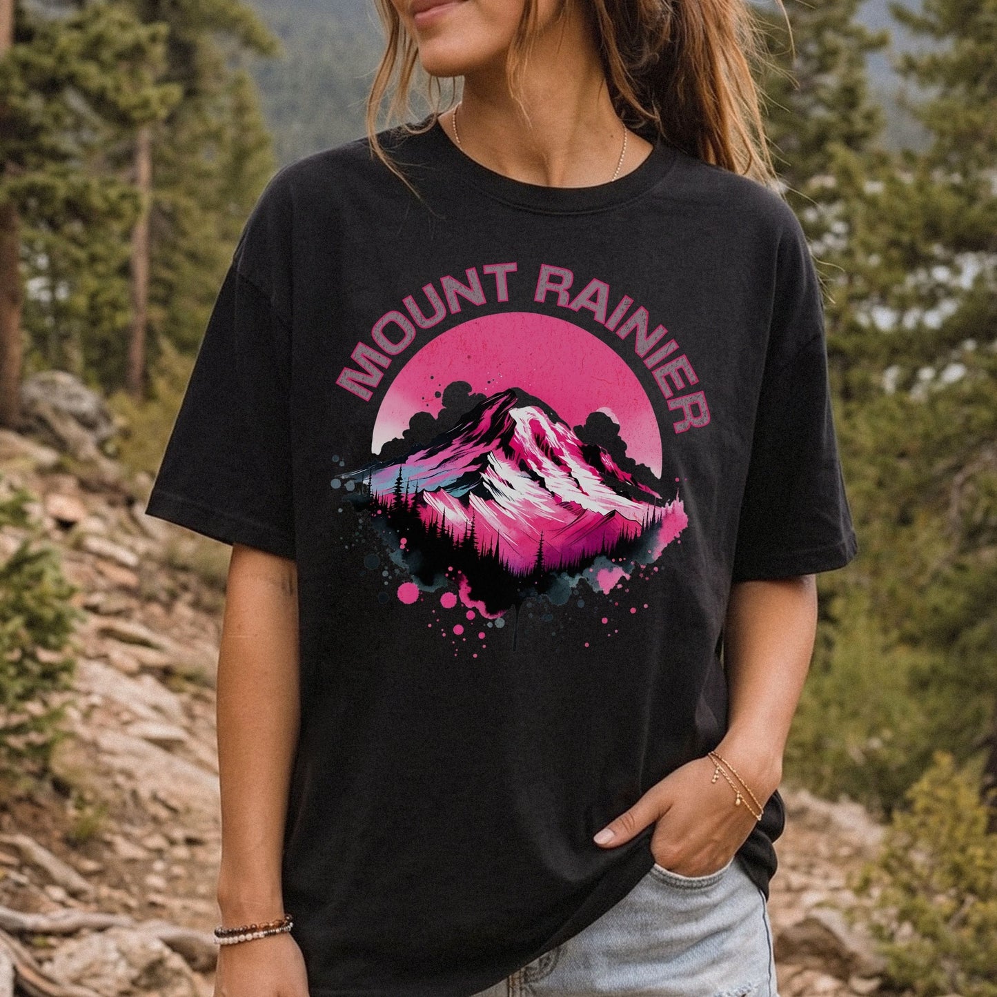 A woman wearing a black graphic t-shirt with a pink and white mountain design and the text 'MOUNT RAINIER' on the front, set against a blurred outdoor mountain forest background.