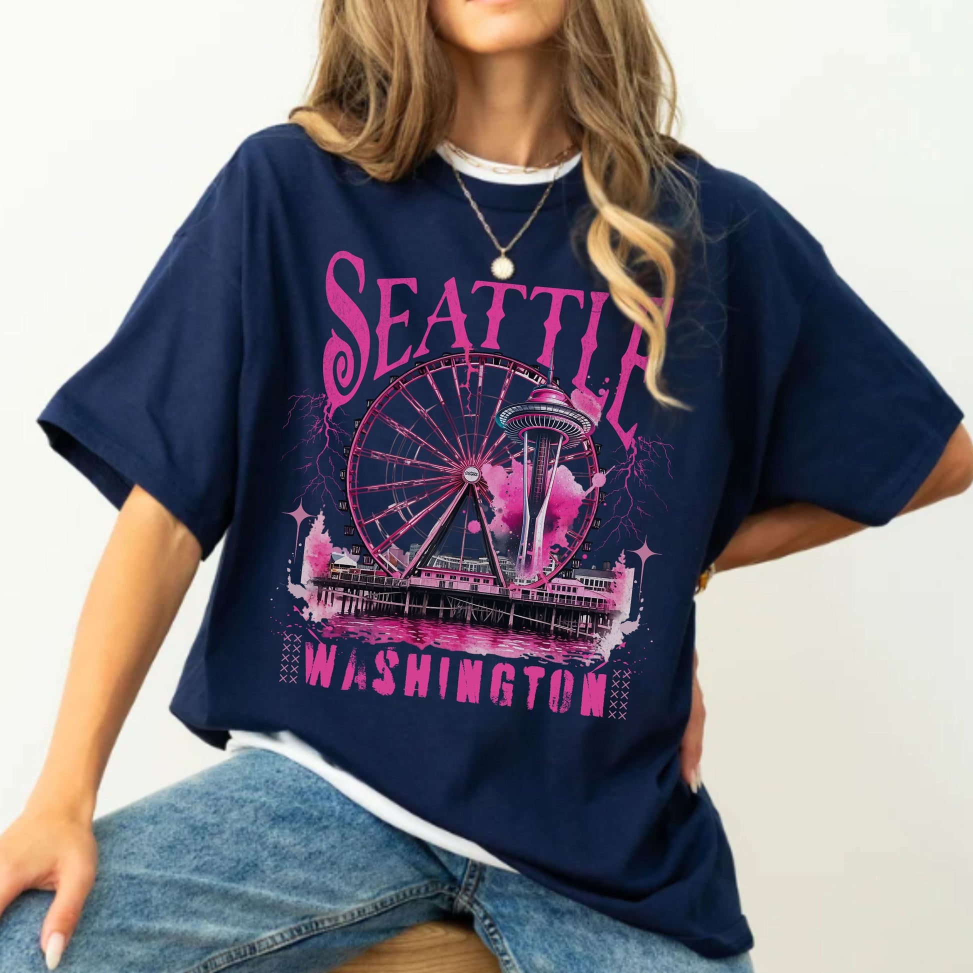 A woman wearing an oversized navy blue graphic T-shirt with a pink and white Seattle, Washington print featuring the Space Needle and Ferris wheel, paired with blue jeans against a plain light background.