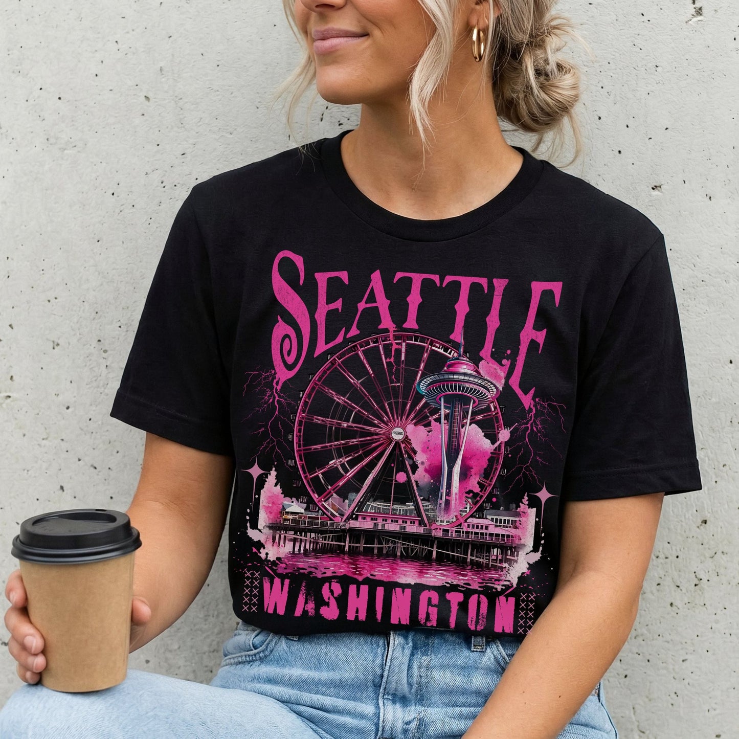A woman wearing a black graphic t-shirt with a pink Seattle, Washington Ferris wheel print, holding a coffee cup against a concrete wall.