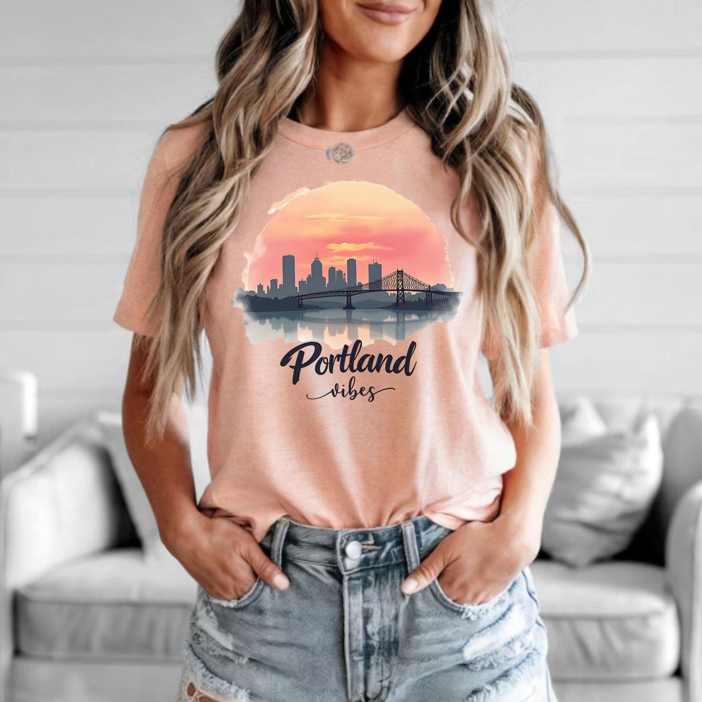 A woman wearing a peach-colored graphic t-shirt with a Portland skyline and sunset print, paired with distressed denim shorts, standing in a living room setting.