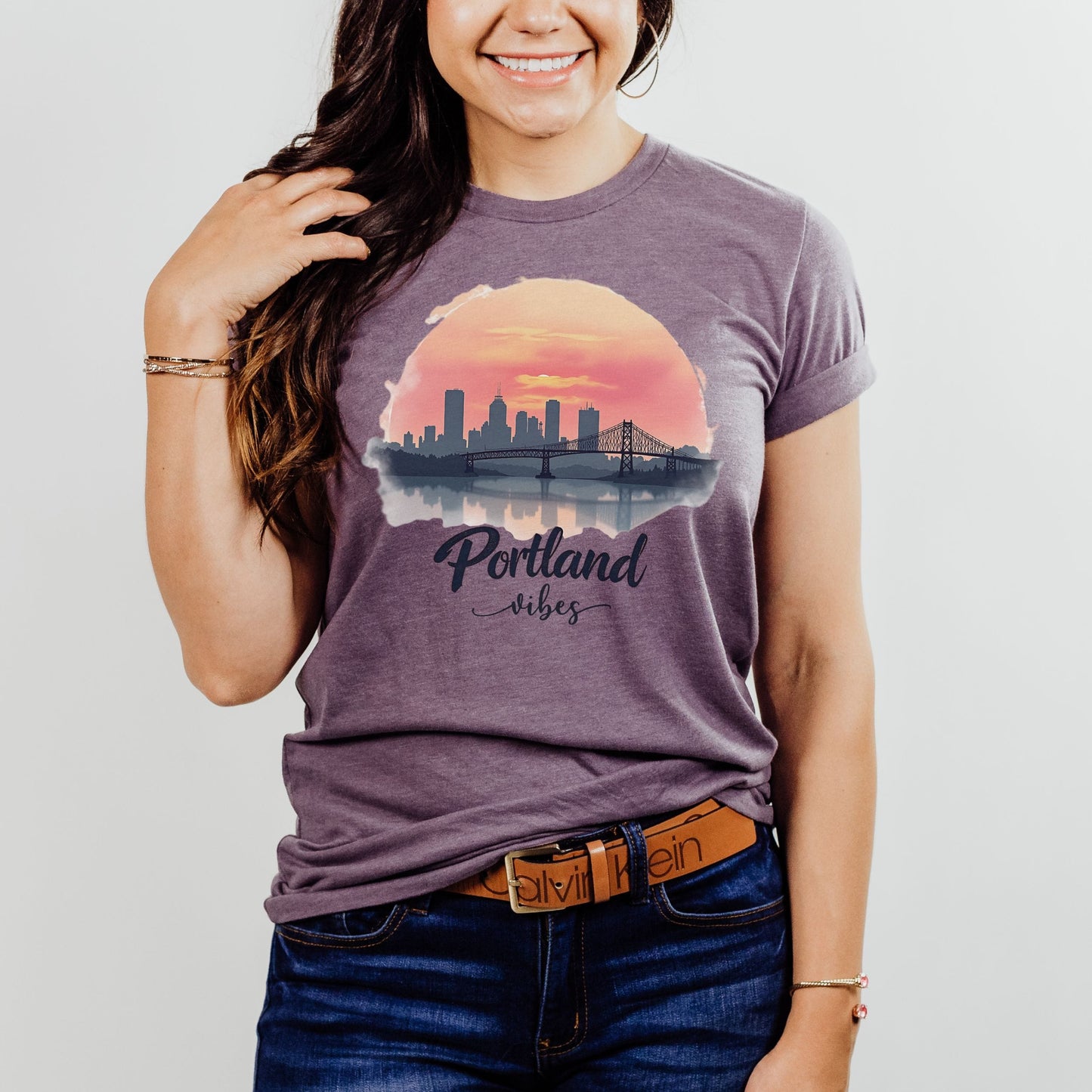 A woman wearing a heathered purple graphic t-shirt with a Portland city skyline and sunset print, paired with blue jeans and a brown belt, against a light gray background.