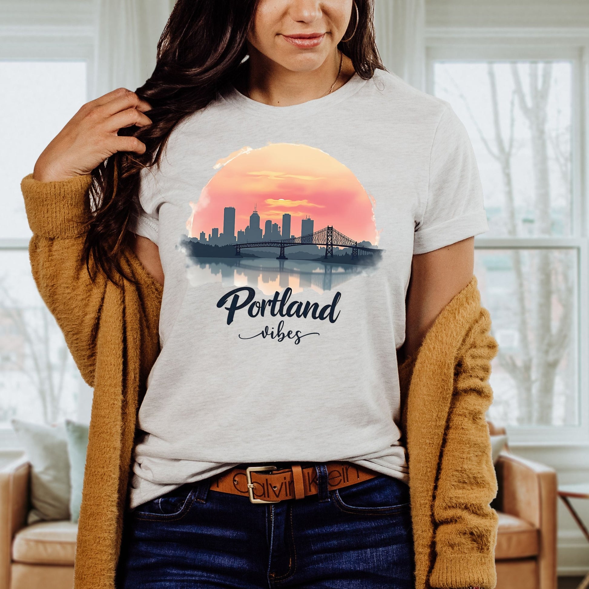 A woman wearing a light gray heather t-shirt with a 'Portland vibes' sunset and skyline graphic, styled with a mustard cardigan and jeans in a bright living room setting.