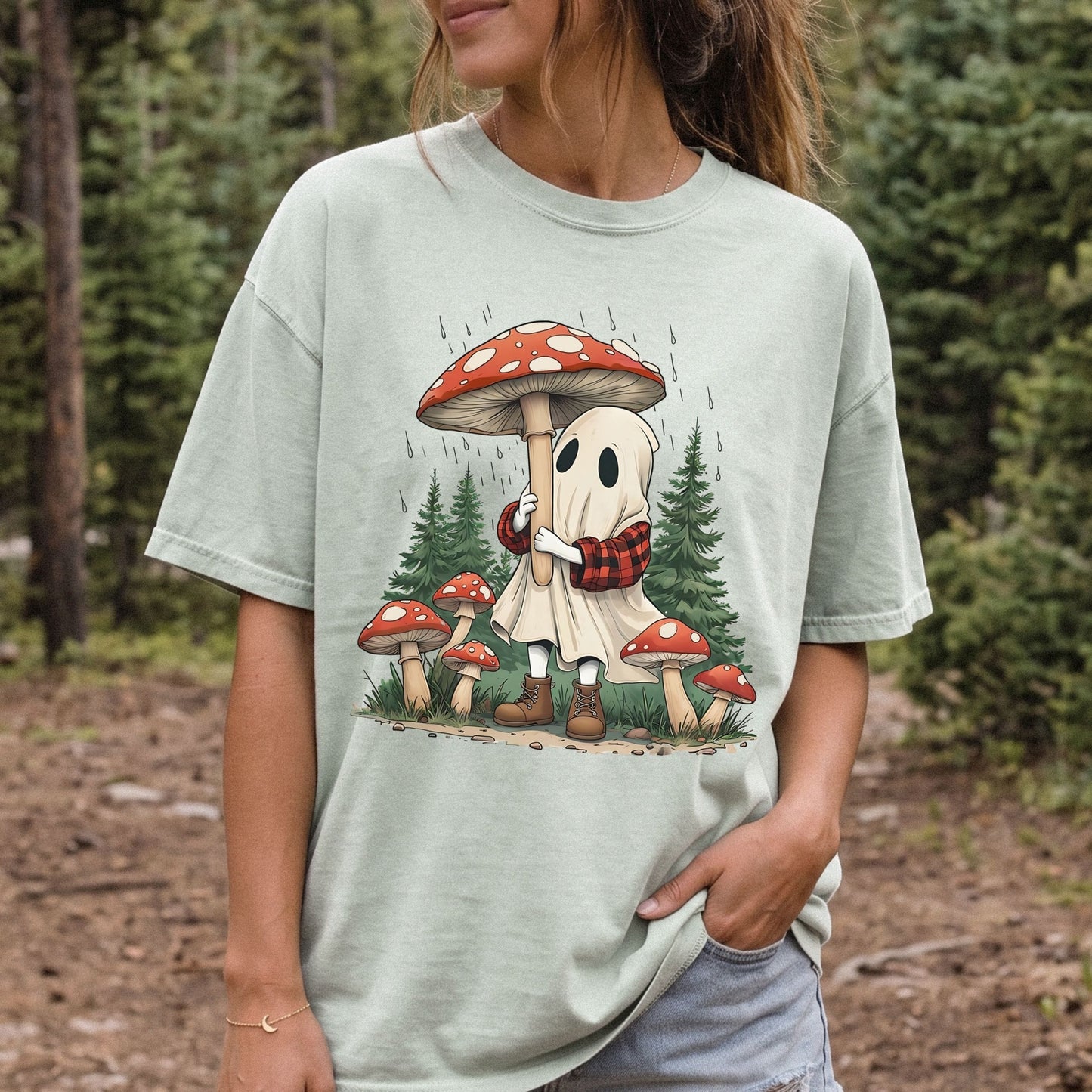 A woman wearing a light green graphic t-shirt in an outdoor forest setting. The t-shirt features a cartoon illustration of a cute white ghost wearing a red and black plaid scarf and brown boots, hugging a large red and white spotted mushroom, with smaller mushrooms and pine trees in the background.