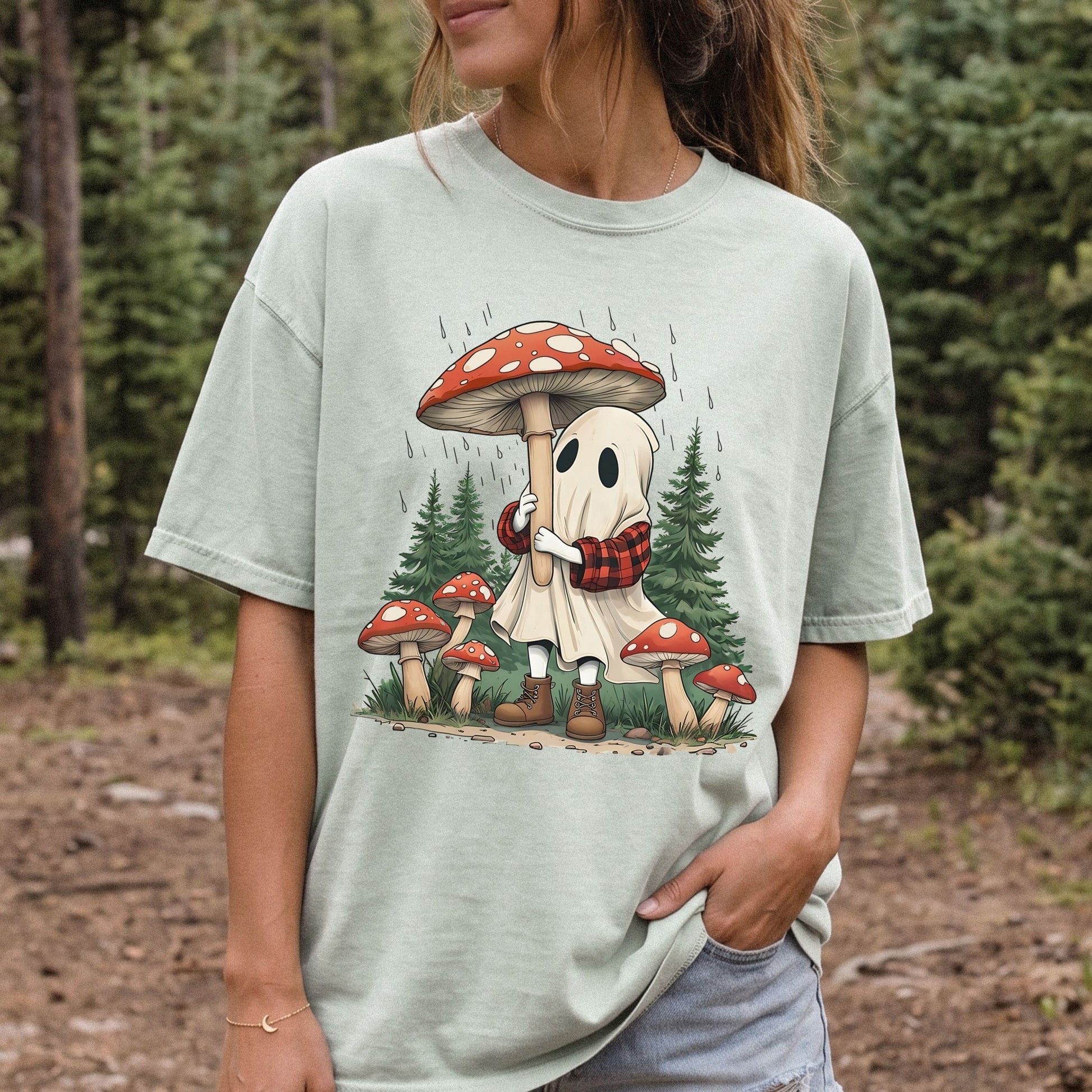 A woman wearing a light green graphic t-shirt in an outdoor forest setting. The t-shirt features a cartoon illustration of a cute white ghost wearing a red and black plaid scarf and brown boots, hugging a large red and white spotted mushroom, with smaller mushrooms and pine trees in the background.