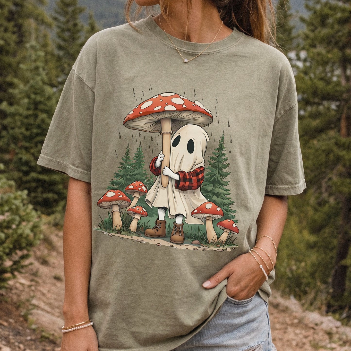 A woman wearing an oversized khaki colored  graphic t-shirt with a cartoon ghost character holding a red and white mushroom in a forest setting.