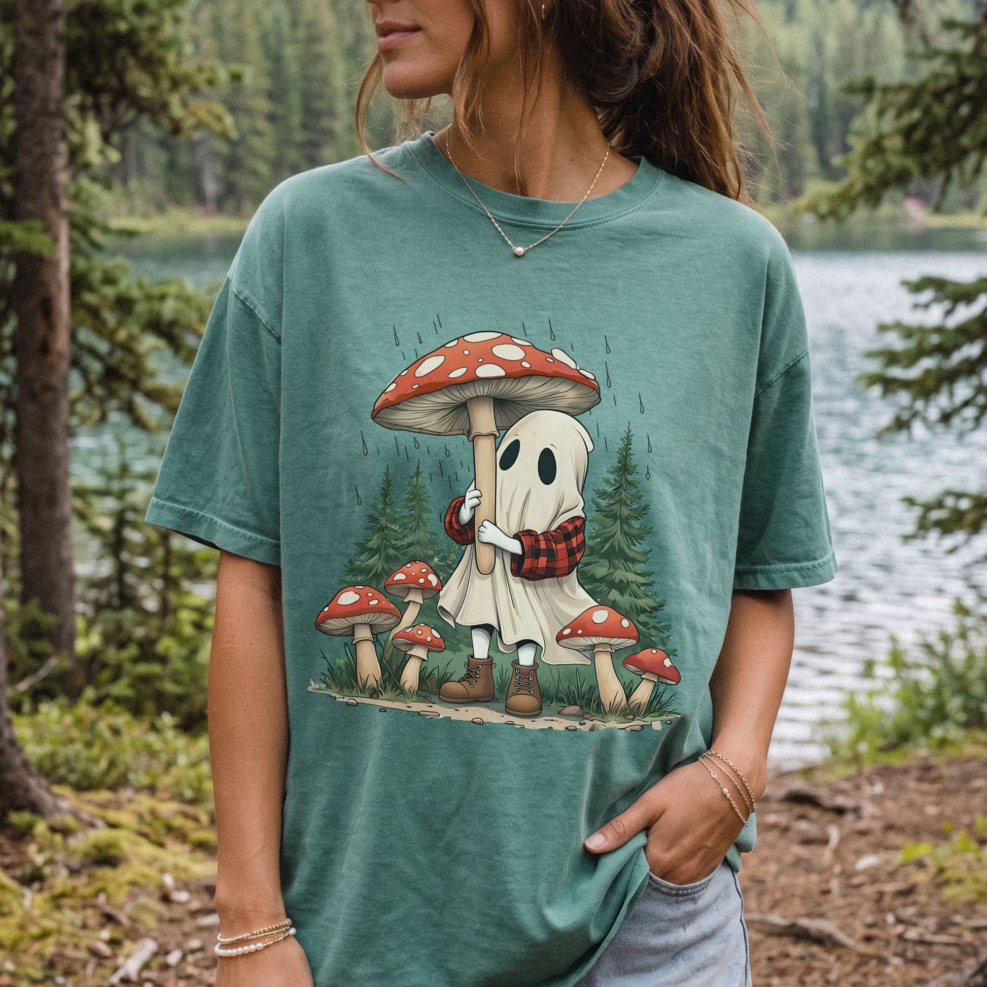 A woman wearing a light green Comfort Colors t-shirt with a cute ghost character under a large red and white mushroom, set against a forest and lake background.