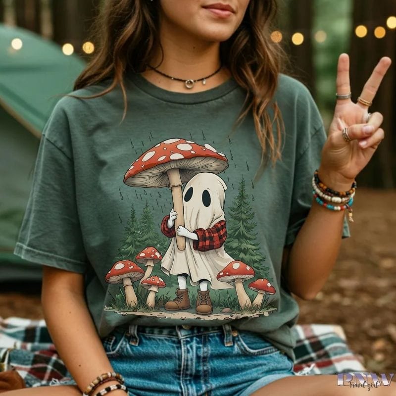 Person wearing a moss green t-shirt with a ghost and mushroom design, sitting outdoors.
