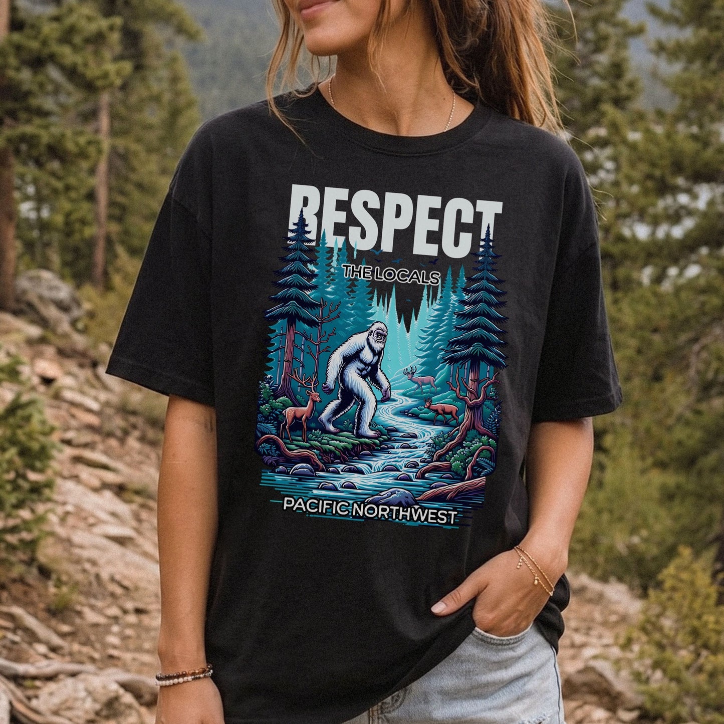 A woman wearing a black graphic t-shirt in an outdoor forest setting. The t-shirt features a colorful graphic of a white yeti in a forest stream with the text 'RESPECT THE Locals' and 'PACIFIC NORTHWEST'.