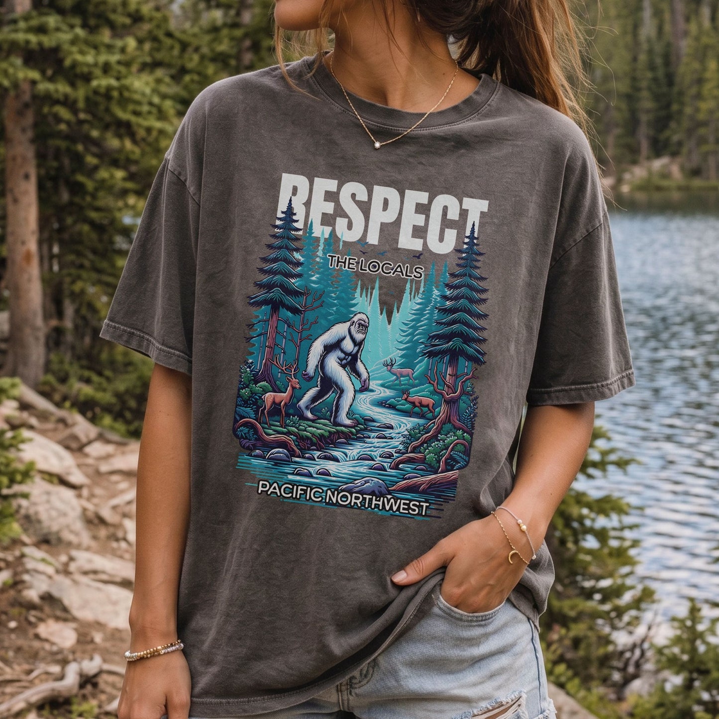 A woman wearing a dark gray mineral-wash graphic t-shirt with a 'Respect the Locals' butthead graphic in a forest setting next to a lake.