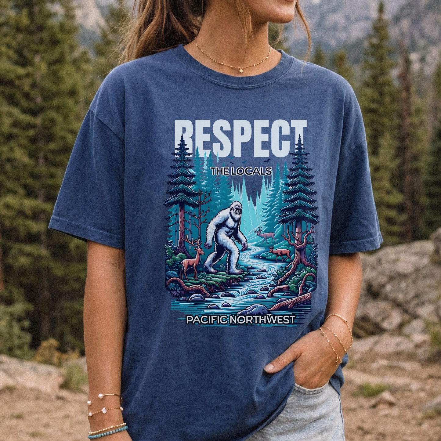 A woman wearing a navy blue graphic t-shirt in an outdoor mountain pine forest setting. The shirt features a colorful illustration of a Bigfoot figure by a stream with the text 'RESPECT THE LOCALS' and 'PACIFIC NORTHWEST'.