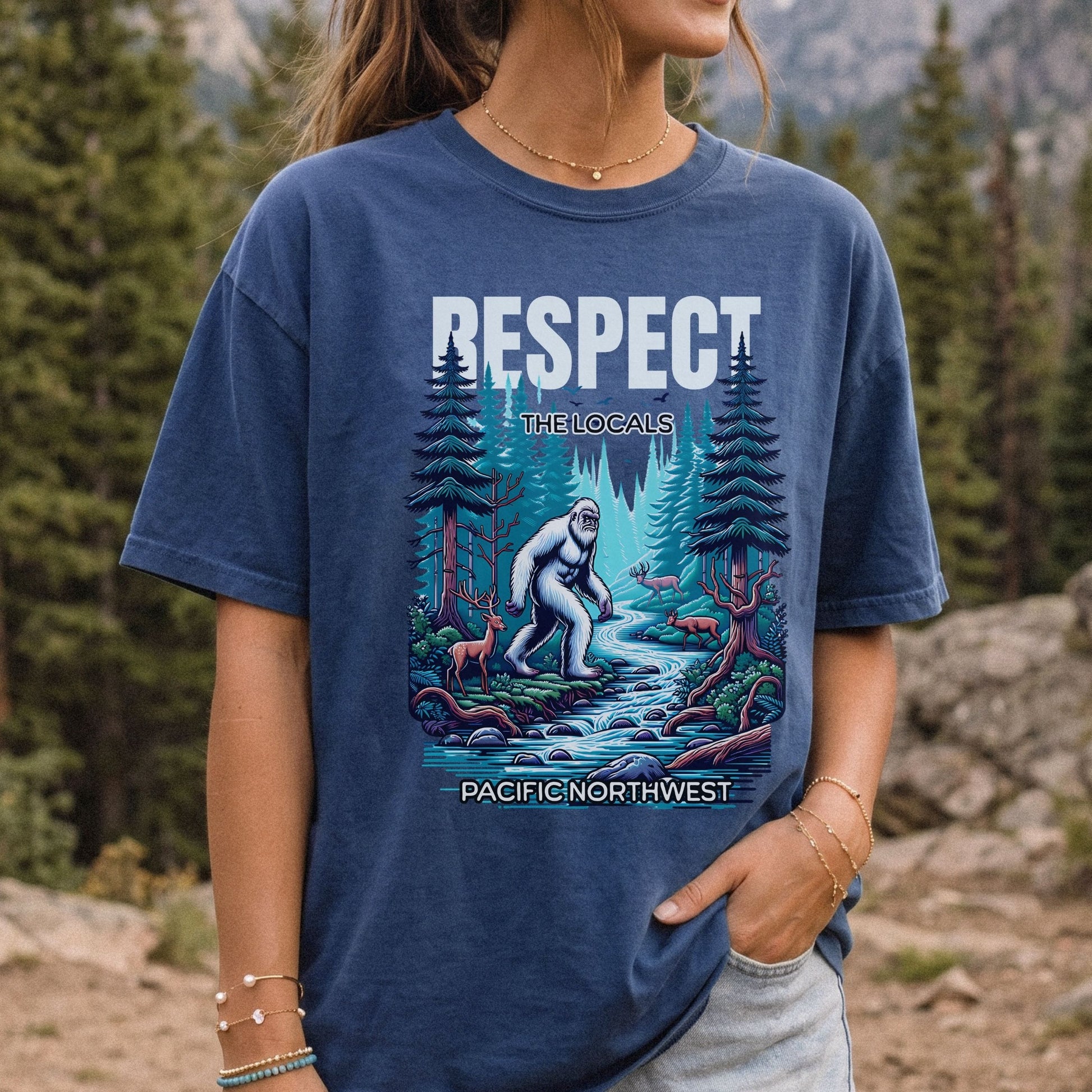 A woman wearing a navy blue graphic t-shirt in an outdoor mountain pine forest setting. The shirt features a colorful illustration of a Bigfoot figure by a stream with the text 'RESPECT THE LOCALS' and 'PACIFIC NORTHWEST'.