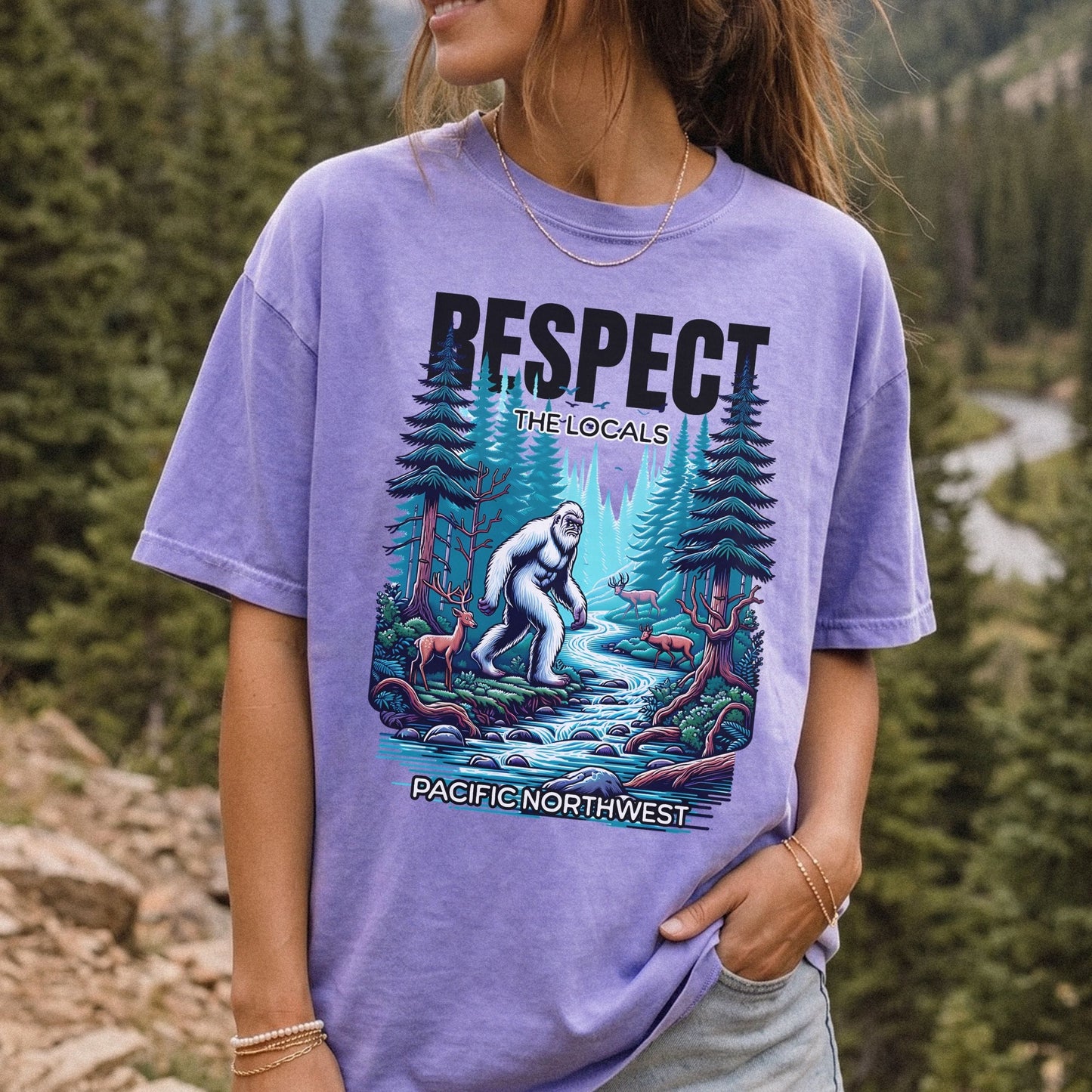 A woman wearing a lavender graphic t-shirt with a 'Respect the Locals' Bigfoot design, set against a blurred forest background.