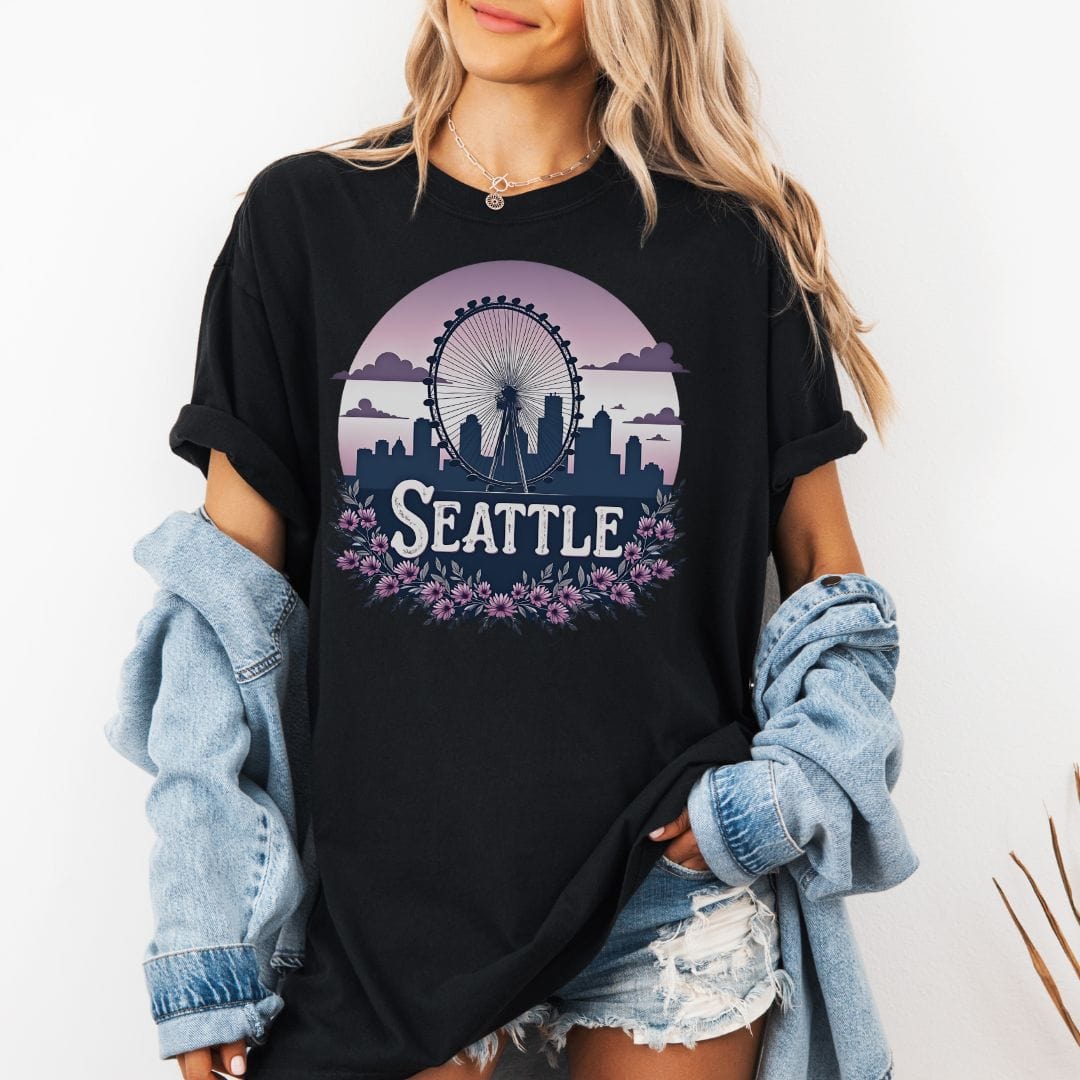 Woman wearing an oversized black graphic t-shirt featuring a retro circular design of the Seattle Great Wheel and city skyline against a pastel pink gradient sky with a purple floral border.