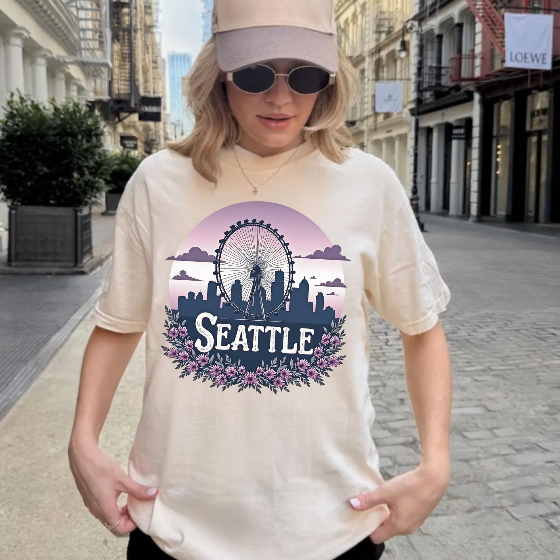A woman wearing a beige baseball cap, sunglasses, and a cream-colored t-shirt with a colorful Seattle graphic print, standing on a city street.