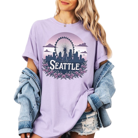 Woman wearing an oversized light purple graphic t-shirt featuring a retro circular design of the Seattle Great Wheel and city skyline against a pastel pink gradient sky with a purple floral border.