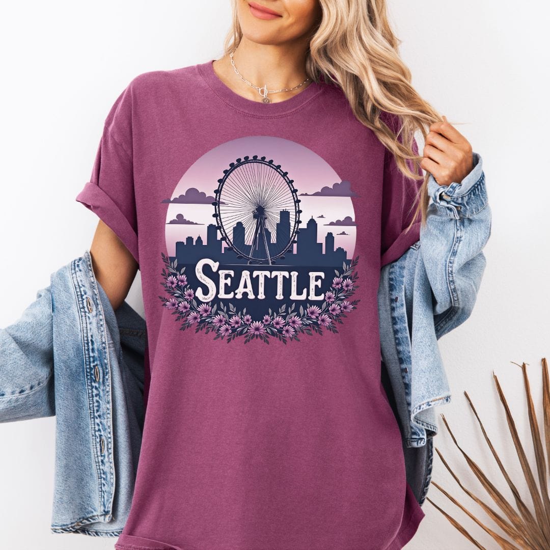 Woman wearing an oversized  purple graphic t-shirt featuring a retro circular design of the Seattle Great Wheel and city skyline against a pastel pink gradient sky with a purple floral border.