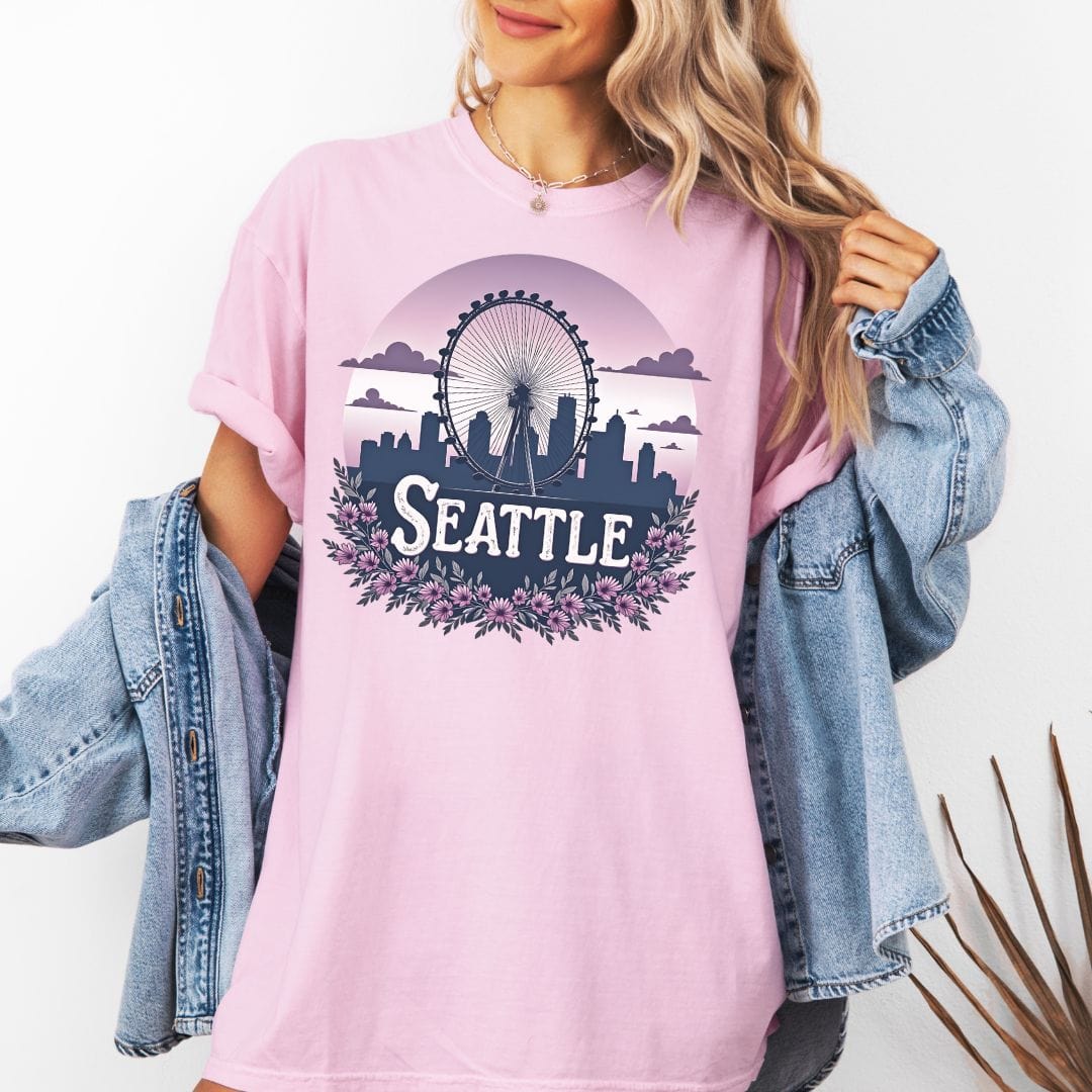 Woman wearing an oversized pink graphic t-shirt featuring a retro circular design of the Seattle Great Wheel and city skyline against a pastel pink gradient sky with a purple floral border.
