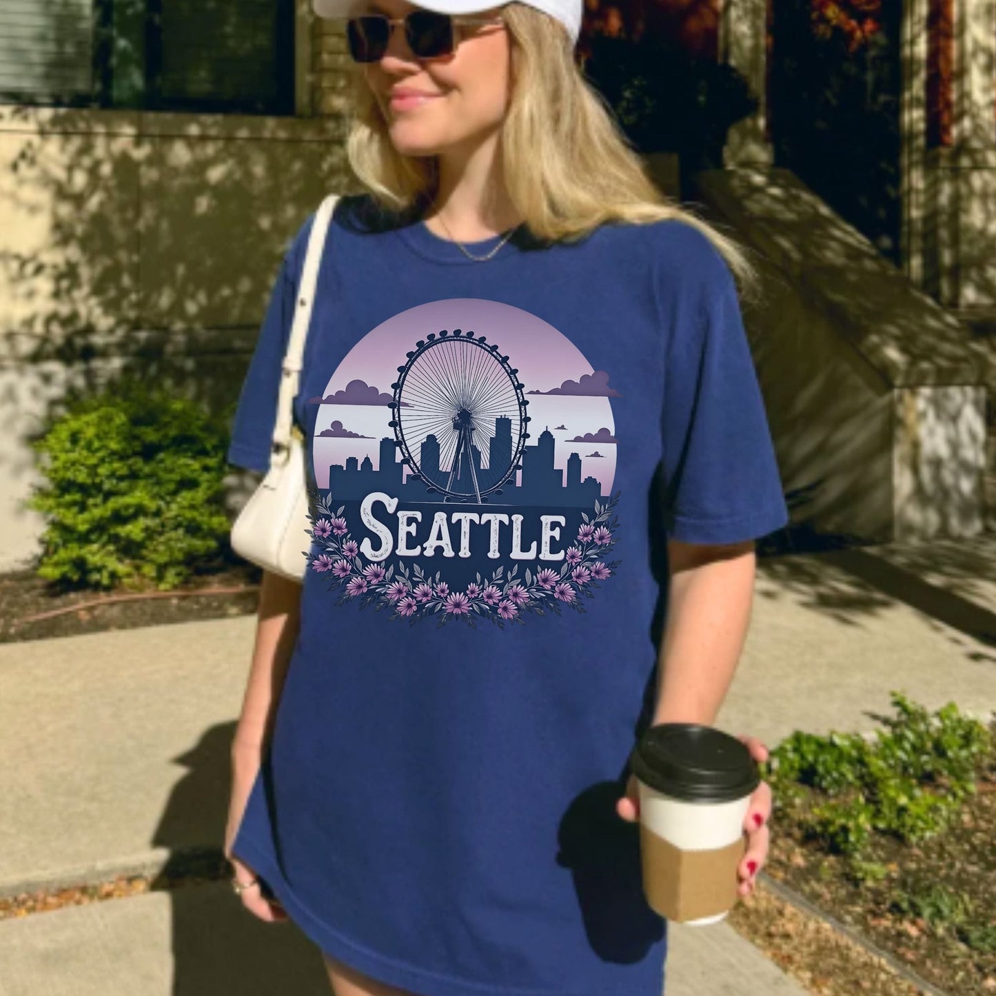 A woman wearing an oversized blue graphic T-shirt with a Seattle skyline and Ferris wheel design, holding a coffee cup outdoors on a sunny sidewalk.