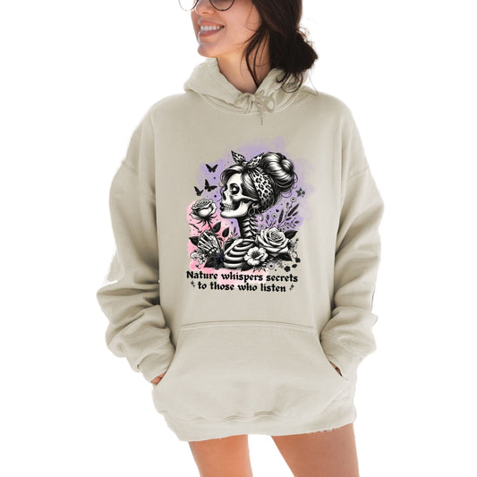 A sand colored hooded sweatshirt with text and a graphic design featuring a skull, flowers, and the phrase 'Nature Whispers, Softstyle Hoodie' on the front.