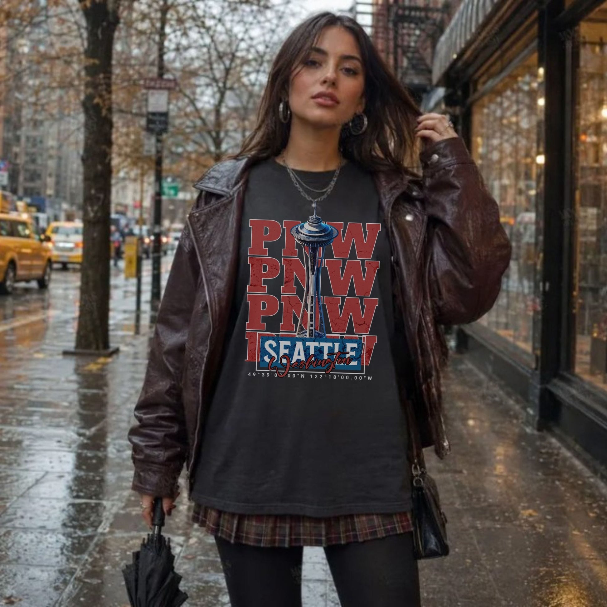 A woman stands on a wet city street wearing a dark graphic T-shirt with stacked red and blue 'PNW' text and a 'Seattle' logo, layered under a brown leather jacket and a plaid shirt underneath.