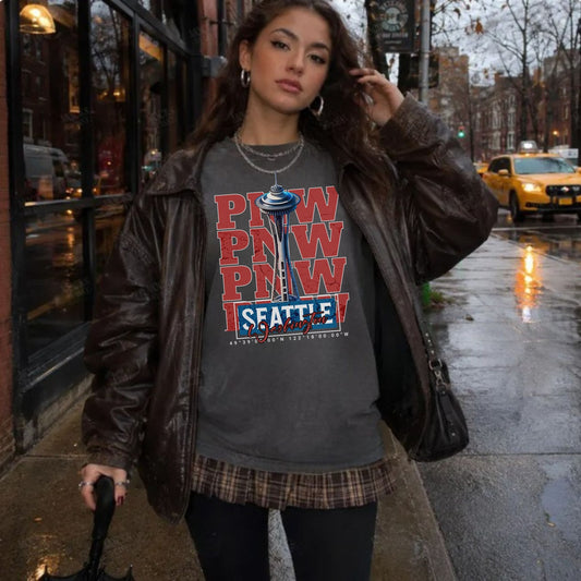 A woman stands on a wet city sidewalk wearing a dark brown faux-leather jacket over a gray graphic T-shirt with red and blue 'PNW SEATTLE' text and a plaid skirt.