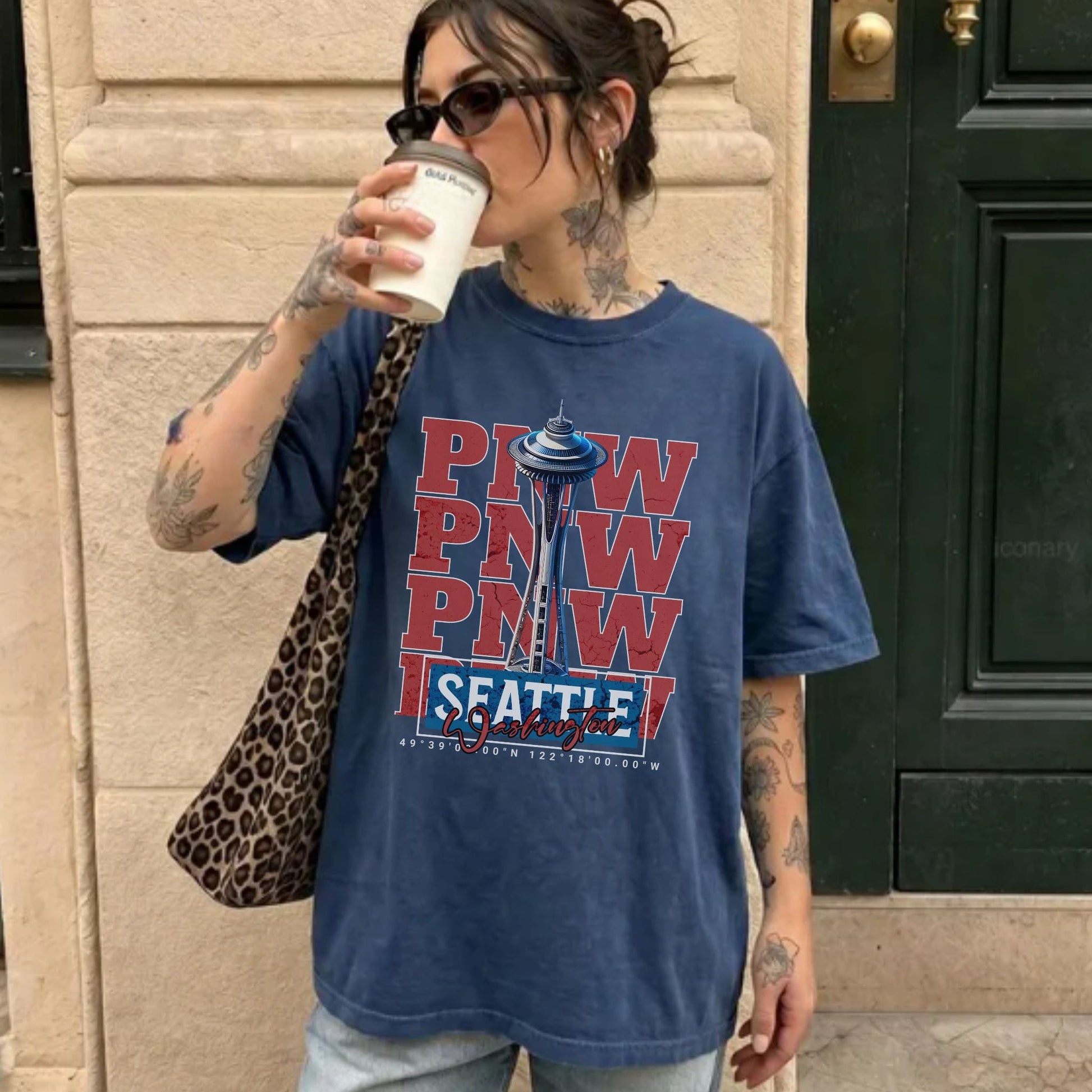 A woman wearing a blue graphic t-shirt with 'PNW, SEATTLE' and the Space Needle printed on the front, holding a coffee cup and wearing a leopard print shoulder bag in front of a beige stone wall and dark green door.