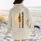 Person wearing a beige hoodie with a tree and 'Adventure' design on a beach