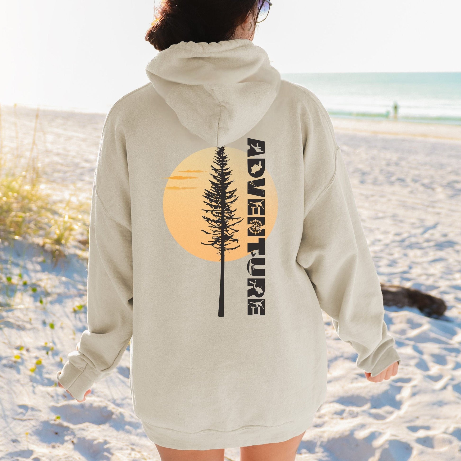 Person wearing a beige hoodie with a tree and 'Adventure' design on a beach