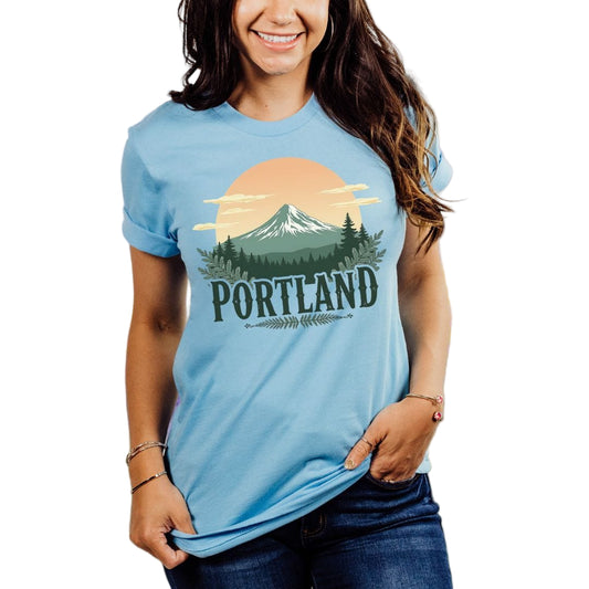 A person wearing a blue t-shirt with a graphic print of a sunrise mountain scene and the word 'Portland' on it.