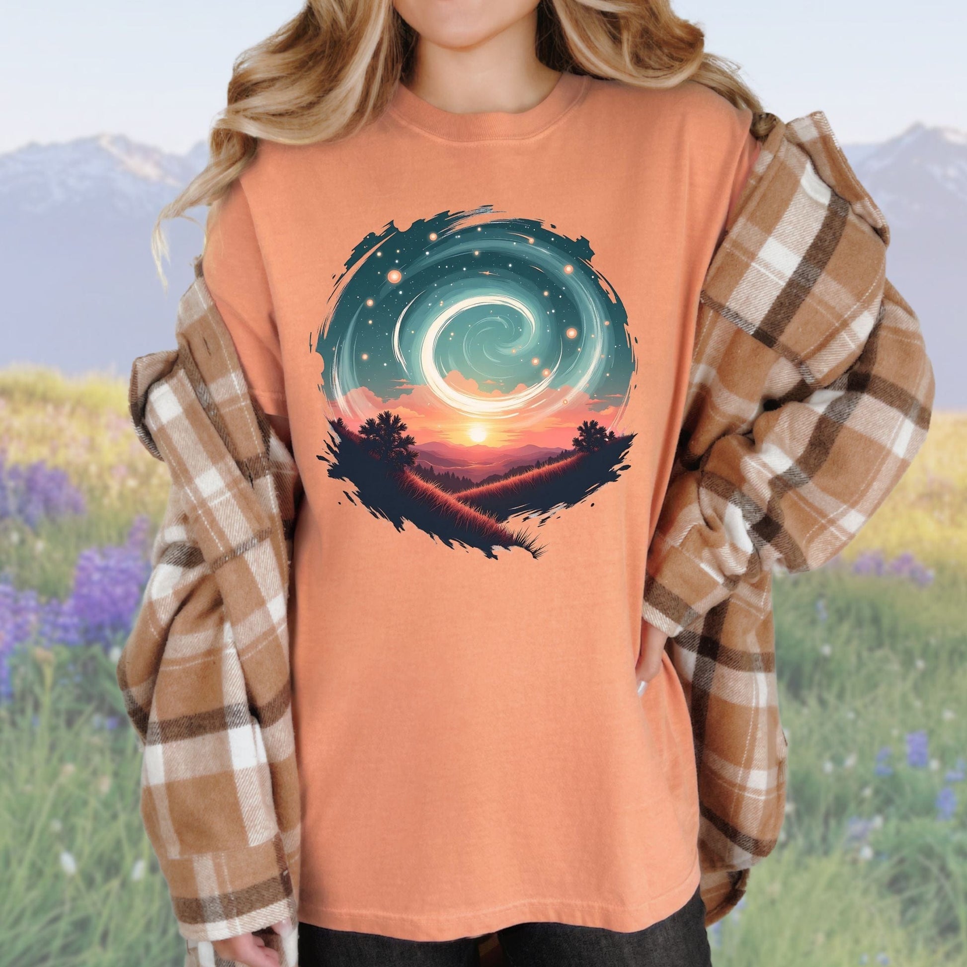 A person wearing a vintage-style graphic tee in a terracotta color with a swirl pattern in the design, standing in a field with mountains in the background.