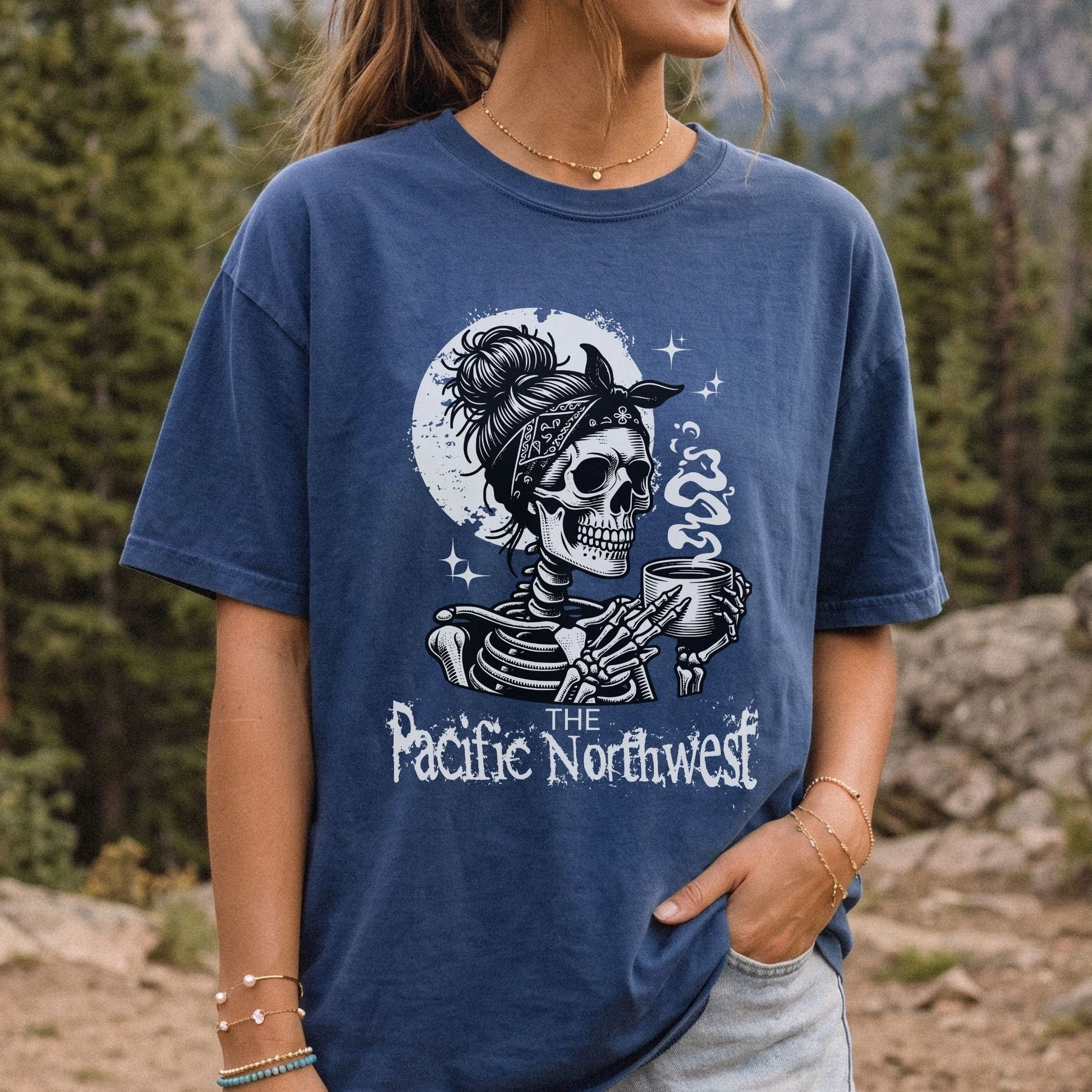 A woman wearing a navy blue graphic t-shirt with a white and gray illustration of a skeleton holding a mug, with the text 'The Pacific Northwest' and 'SOS'. The background is a blurred outdoor mountain and pine forest environment.