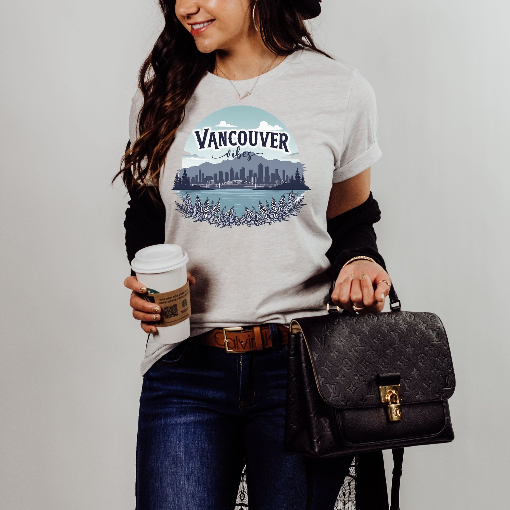 A woman wearing a light gray graphic t-shirt with a 'Vancouver Blues' band logo and skyline print, styled with a black long-sleeve shirt underneath, dark blue jeans, and holding a black monogram handbag and a coffee cup.
