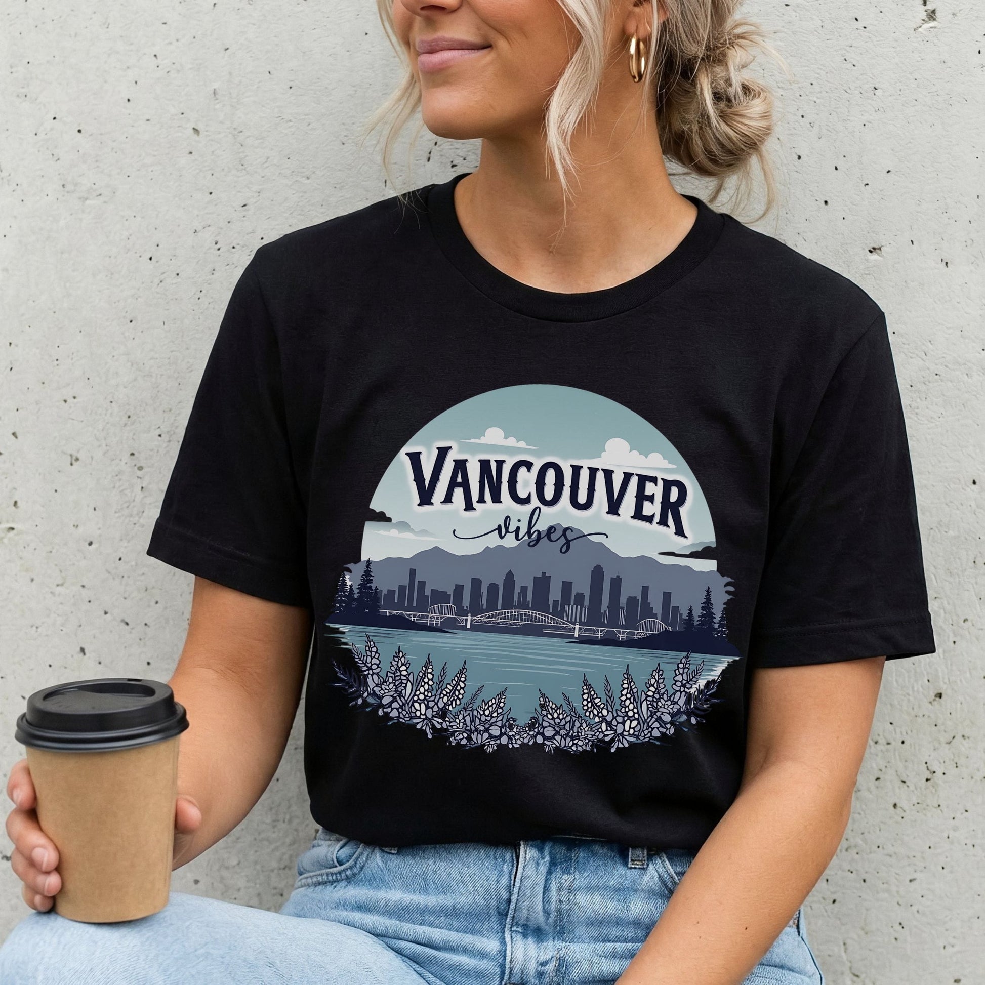 A woman wearing a black graphic t-shirt with a 'Vancouver Vibes' cityscape print, holding a coffee cup against a concrete wall.