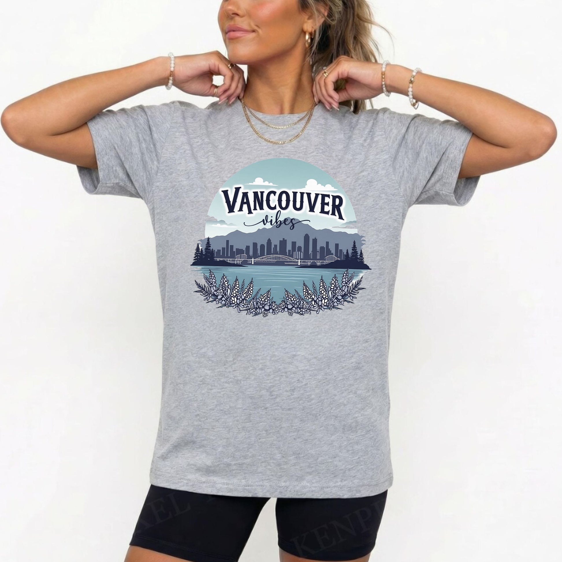 A woman wearing a heather gray graphic t-shirt with a 'Vancouver Vibes' print featuring a city skyline, mountains, and water, paired with black shorts against a plain white background.