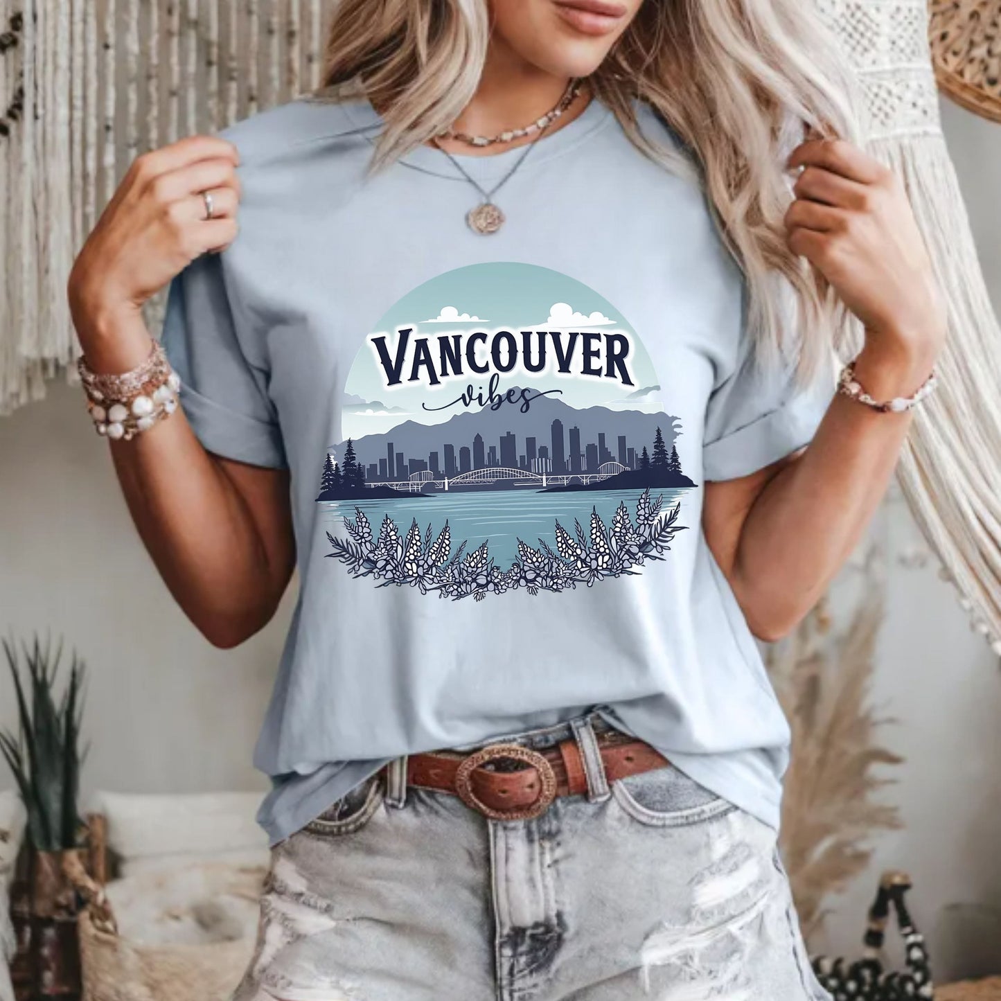 A woman wearing a light blue graphic t-shirt with a 'Vancouver Vibes' cityscape print, paired with distressed light-wash jeans and jewelry, in a bohemian-style indoor setting.