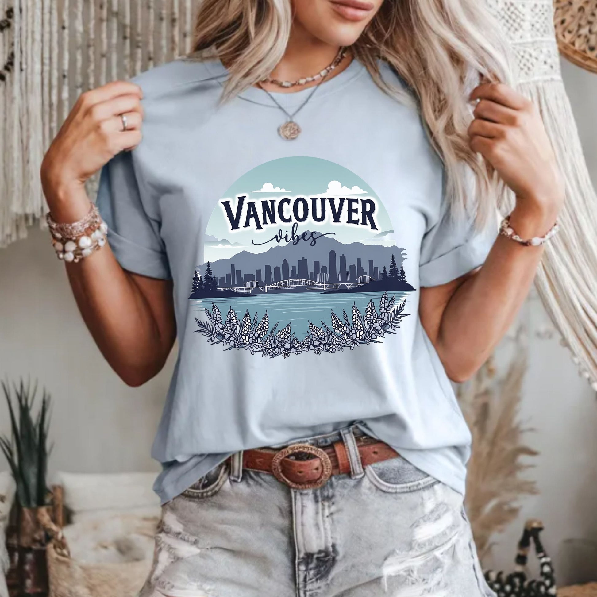 A woman wearing a light blue graphic t-shirt with a 'Vancouver Vibes' cityscape print, paired with distressed light-wash jeans and jewelry, in a bohemian-style indoor setting.