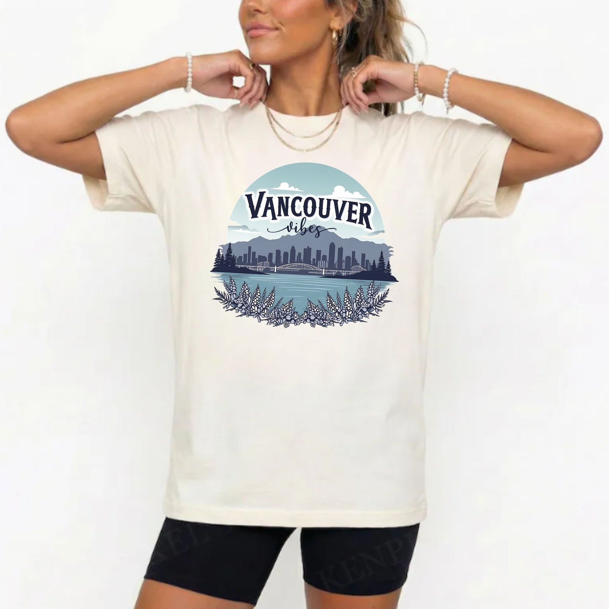 A woman wearing a cream-colored t-shirt with a graphic print of the Vancouver skyline and lake under a blue sky. She is also wearing black bike shorts and gold jewelry, posing against a plain white background.