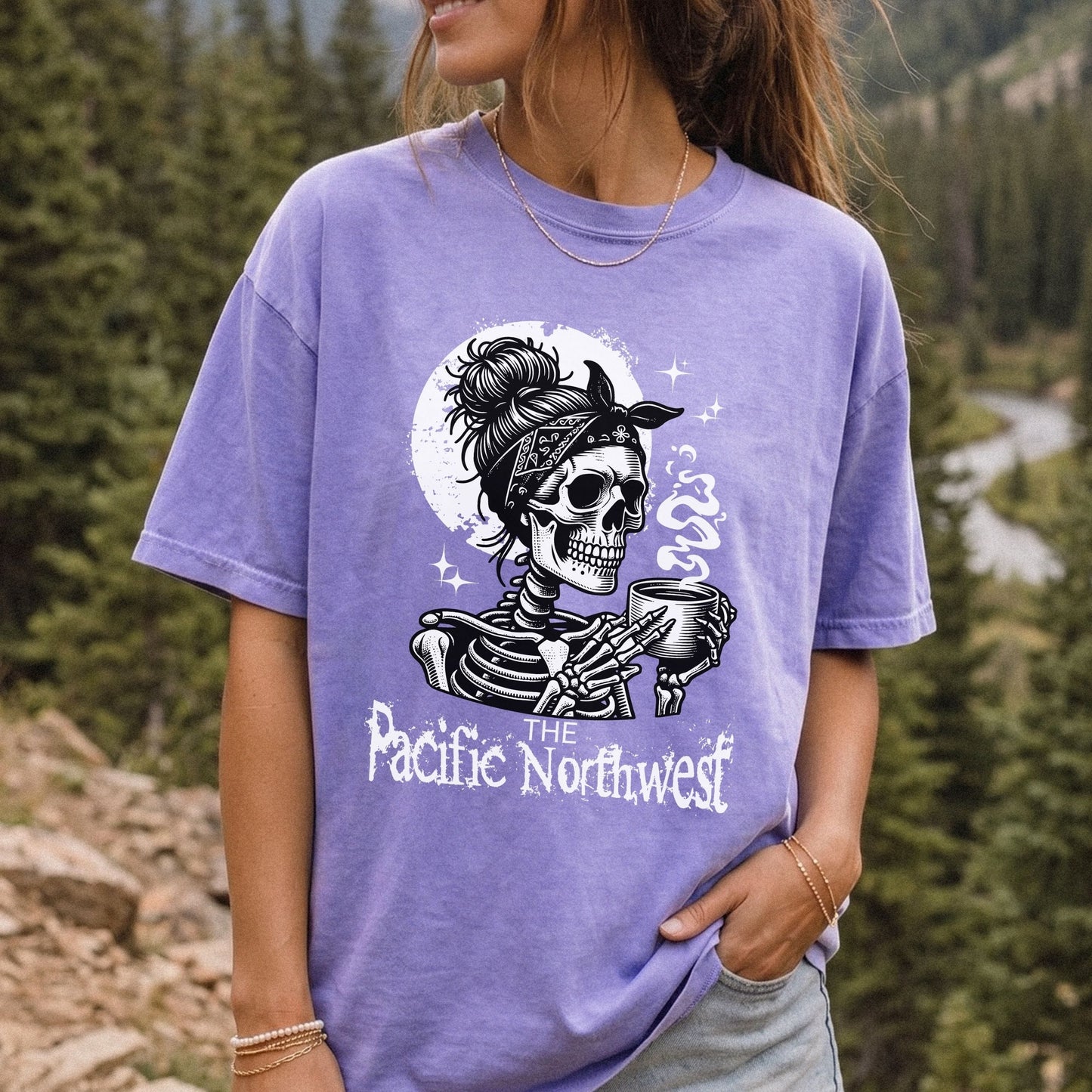 A woman wearing a lavender purple graphic t-shirt with a black and white skeleton design holding a mug, with the text 'The Pacific Northwest' printed below. The background is a blurred outdoor mountain forest scene.