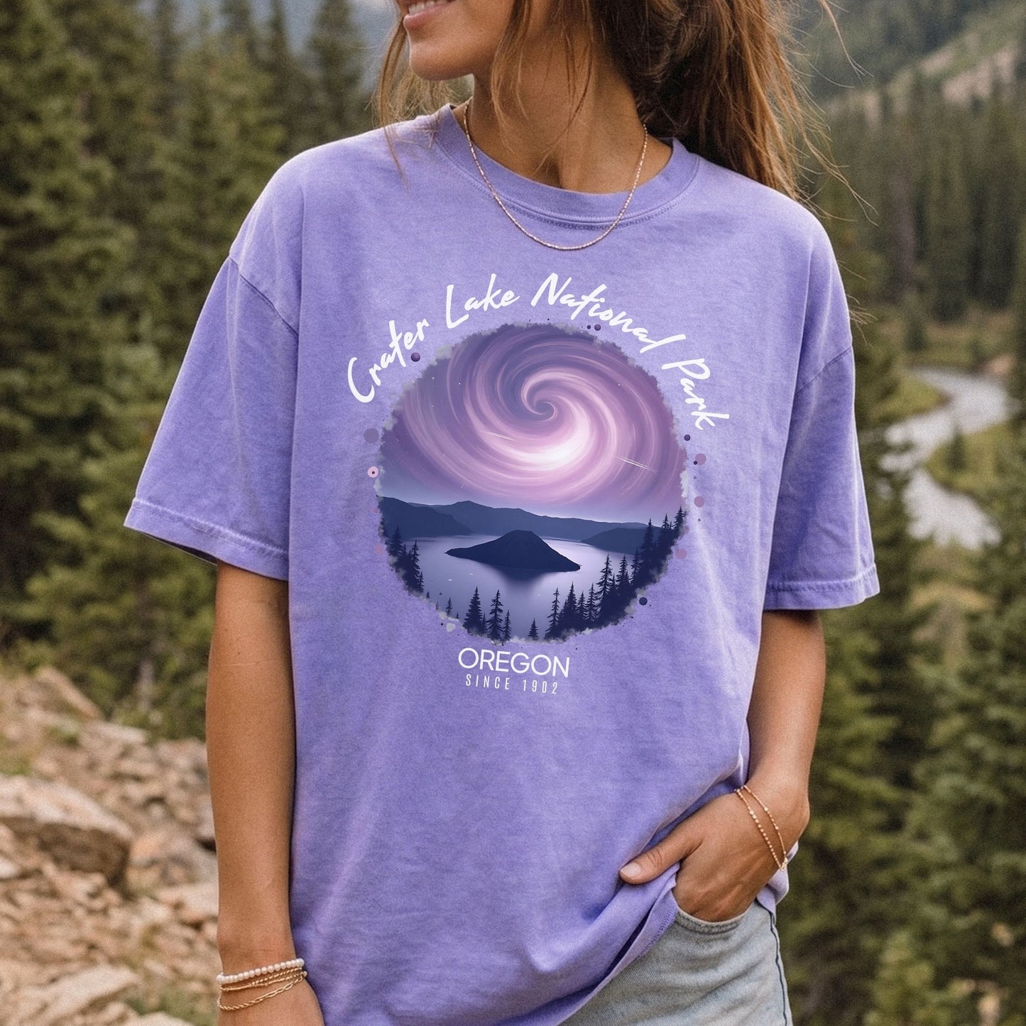 A woman wearing a Comfort Colors violet t-shirt with a vintage-style circular graphic of Crater Lake at night under a purple spiral galaxy, framed by white cursive text that reads Crater Lake National Park Oregon.