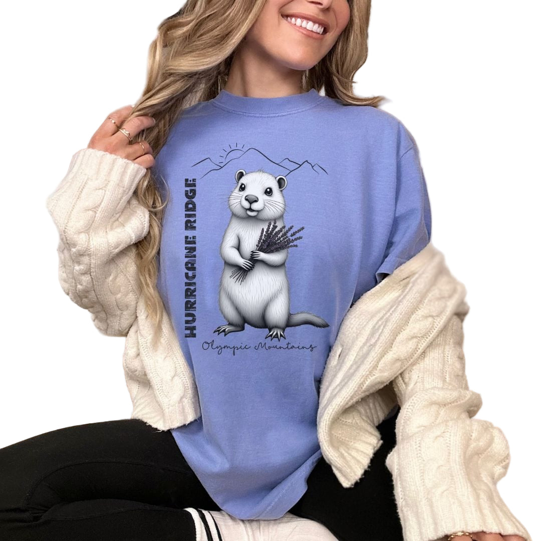A person wearing a blue vintage-style t-shirt with an illustration of a marmot and the text 'Hurricane Ridge'. They are sitting in a cozy room with a white blanket and a cream-colored jacket.