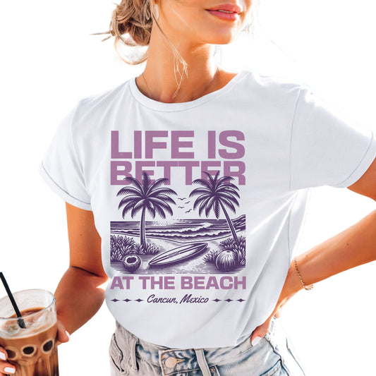 A woman at the beach wearing a white t-shirt with the text 'LIFE IS BETTER AT THE BEACH' and a graphic of palm trees. She is holding a beverage and smiling.